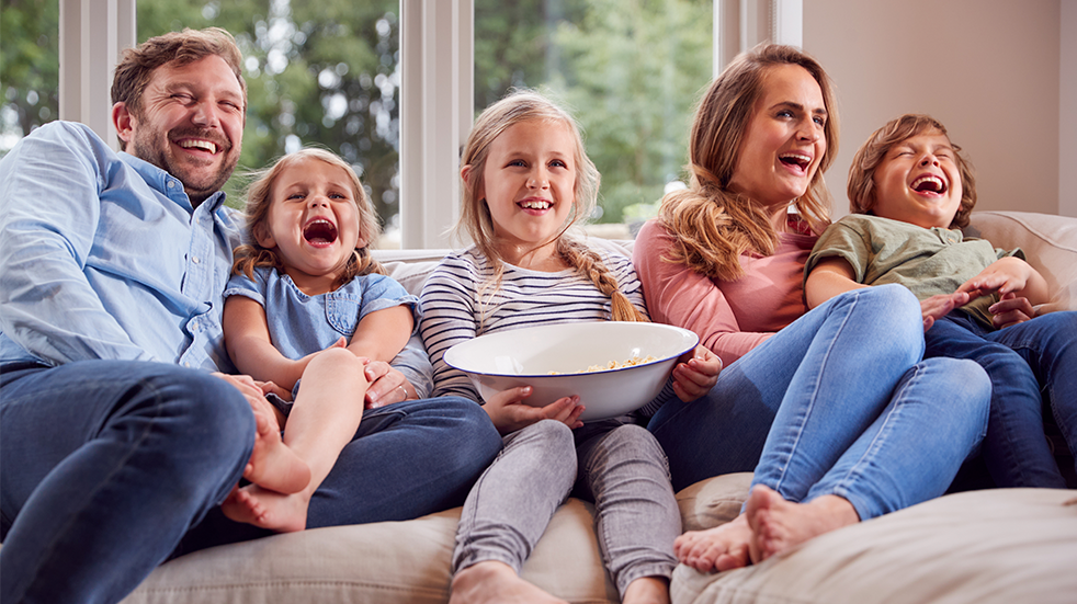 A family of five sitting on sofa looking happy watching a film with a bowl of popcorn